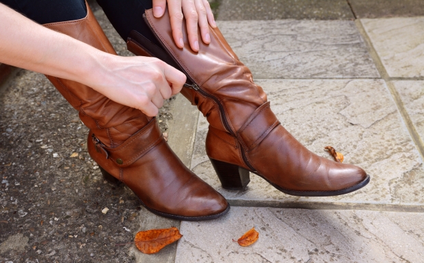 Woman zips up high-heeled tan leather boots, autumn leaves scattered at her feet